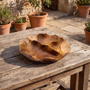 Può includere: Una grande ciotola di legno intagliata a mano con una forma ondulata e organica. La ciotola è in legno marrone chiaro e poggia su un tavolo di legno invecchiato. Lo sfondo include vasi di terracotta con piante.