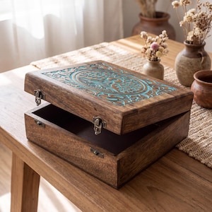 May include: A rectangular wooden box with a hinged lid featuring a carved circular tree design highlighted in teal. The box has a dark brown finish, a smooth texture, and is secured with two small metal latches on the front edge of the lid and base.