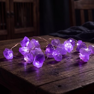 Op de afbeelding: Een LED-lichtsnoer met ruwe, doorschijnende paarse amethistkristallen bevestigd aan een dunne koperdraad. De kristallen gloeien met een levendig violet licht en vormen een lichtgevend geheel van grillige, natuurlijke steenvormen.