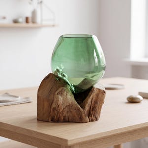 Peut inclure: Un vase en verre vert de forme arrondie, posé sur un socle en bois naturel. Le vase a une surface lisse et une légère teinte verte. Le socle en bois est taillé grossièrement, révélant le grain et la texture naturels du bois.