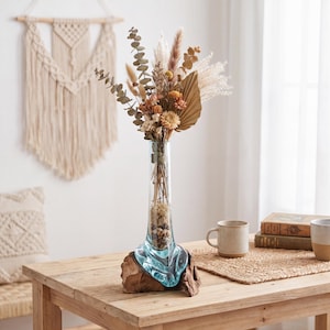 Könnte beinhalten: Eine klare Glasvase, die an einen Sockel aus natürlichem Teakholz angepasst ist, gefüllt mit einem Strauß getrockneter Blumen, darunter Eukalyptus, Pampasgras und kleine beige Blüten, mit Kieselsteinen am Boden des Glasgefäßes.