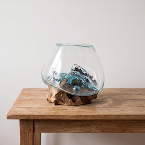 Può includere: Una ciotola in vetro trasparente dalla forma organica fusa con una base in legno naturale color marrone chiaro. Il vetro presenta accenti interni colorati di blu che imitano l'aspetto di onde o formazioni rocciose, creando un recipiente fluido.