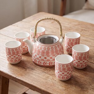 Peut inclure: Un service à thé en céramique composé d'une théière ronde avec une anse en bambou tressé et un infuseur en maille d'acier inoxydable, accompagné de cinq tasses assorties. Chaque pièce présente une base blanche avec un motif floral et géométrique rouge.