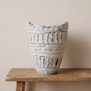 Peut inclure: Un grand vase en céramique blanc cassé avec une surface texturée et une large ouverture incurvée. Le vase présente des bandes horizontales avec des encoches rectangulaires verticales et de petits détails circulaires. Il est posé sur une surface en bois.