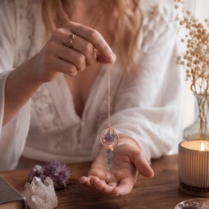 May include: A rose gold pendulum featuring a pointed clear quartz crystal base and a small purple amethyst crystal encased within a circular metal frame, suspended from a delicate rose gold chain held by a hand with a gold ring.