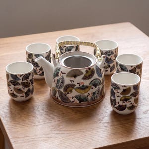 Può includere: Un set da tè in ceramica composto da una teiera con manico in bambù intrecciato e infusore in metallo, accompagnato da cinque tazze abbinate. La superficie in ceramica bianca è decorata con un motivo ripetuto di ventagli pieghevoli colorati nei toni del blu, giallo, marrone e verde.