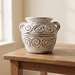 Può includere: Un vaso bianco dall'aspetto invecchiato con due manici e un motivo decorativo a spirale. Il vaso è realizzato in un materiale strutturato e poggia su un tavolo di legno. Il vaso ha una finitura anticata con accenti neri.