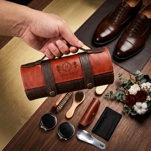 Puede incluir: Un kit de cuidado de calzado cilíndrico de cuero marrón y tostado con asa, que presenta el texto grabado HOWARD y un logotipo de escudo. El kit incluye dos latas negras redondas, un cepillo de madera, un estuche de cuero, un paño y calzador.