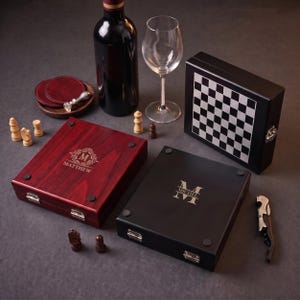 Könnte beinhalten: Zwei Spielkassetten aus Holz, eine rötlich-braune mit dem Namen Matthew und eine schwarze mit dem Namen Michel, dazu eine Weinflasche, ein Glas, ein Korkenzieher, Untersetzer und verstreute Schachfiguren. Eine dritte Schachkassette steht aufrecht.
