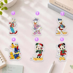 May include: Six acrylic pins featuring Disney characters wrapped in colorful Christmas lights. The characters include Goofy, Daisy Duck, Donald Duck, Pluto, Minnie Mouse, and Mickey Mouse, each numbered one through six in purple circles.