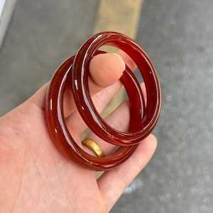 Puede incluir: Dos brazaletes circulares y lisos fabricados con un material rojo translúcido profundo. Los brazaletes presentan un acabado pulido y brillante que refleja la luz, mostrando un tono carmesí rico y consistente en toda su estructura.