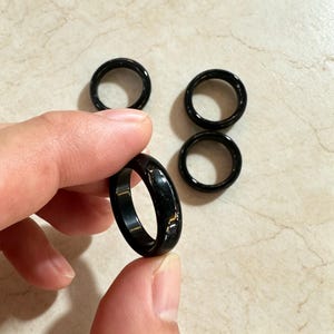 May include: Three smooth, solid black rings with a polished, reflective finish. Each ring features a simple, circular band design with a consistent width and rounded edges, showcasing a deep, uniform black color throughout the entire surface.