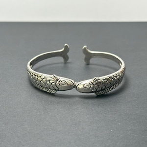 May include: A silver-toned open cuff bracelet featuring two fish heads meeting in the center. The metal surface is textured with detailed scale patterns along the band, and the terminals of the cuff are shaped like small, flat fish tail fins.