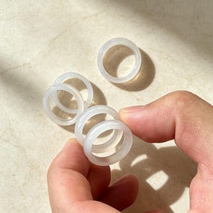Puede incluir: Una colección de cinco anillos blancos translúcidos y lisos con un diseño de banda simple. Los anillos son circulares y presentan un acabado pulido, pareciendo uniformes en color y forma. Estos anillos son ligeros y tienen una superficie lisa.