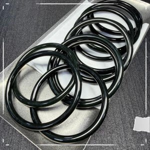Könnte beinhalten: Eine Sammlung dunkelgrüner Jade-Armreifen. Die Armreifen sind rund und poliert, mit glänzendem Finish. Sie sind auf einer weißen Oberfläche angeordnet, was einen Kontrast zur dunklen Farbe des Jades bildet. Die Armreifen sind ein klassisches Schmuckstück.