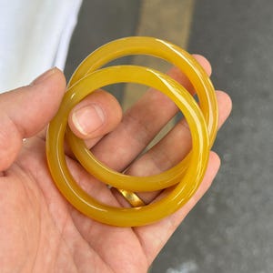 May include: Two smooth, circular bangles crafted from a translucent, mustard-yellow material. The bracelets feature a polished, glossy finish that reflects light, creating a uniform, rounded profile with a consistent thickness throughout each band.