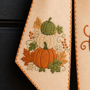 May include: A beige fabric bow with a scalloped orange edge. The left tail features an embroidered stack of three pumpkins in green, white, and orange, surrounded by autumn leaves. The right tail displays a large, embroidered orange letter C.