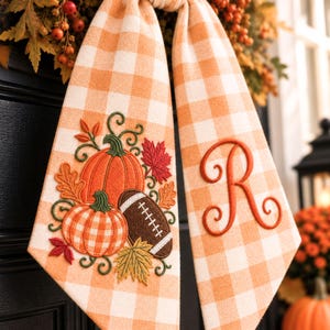 May include: A hanging fabric door decoration featuring an orange and white gingham pattern. One side displays embroidered pumpkins, a football, and autumn leaves. The other side features a large, embroidered, cursive letter R in orange thread.