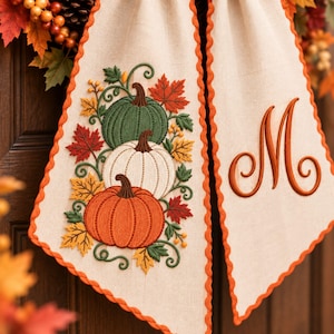 Peut inclure: Une bannière en tissu beige avec une bordure orange festonnée, ornée d'une broderie représentant trois citrouilles verte, blanche et orange, entourées de feuilles d'automne et de vignes, avec une grande lettre M orange brodée à droite.