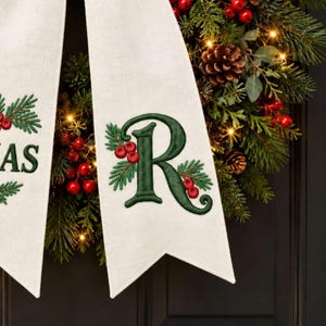 May include: A cream-colored linen ribbon featuring an embroidered dark green letter R, accented with small red berries and green pine needles. The ribbon is draped over a lush evergreen wreath adorned with pinecones and warm white fairy lights.