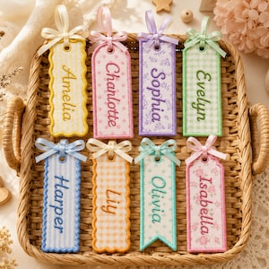 May include: Eight rectangular fabric bookmarks with scalloped or straight edges, each featuring a name embroidered in a matching thread color. The bookmarks display various patterns including stripes, polka dots, gingham, and floral prints, topped with satin ribbons.