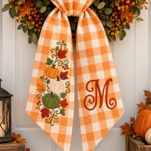 May include: A hanging fabric decoration featuring an orange and white buffalo check pattern. One side displays embroidered pumpkins in orange, white, and green with autumn leaves and berries. The other side features a large, embroidered letter M.