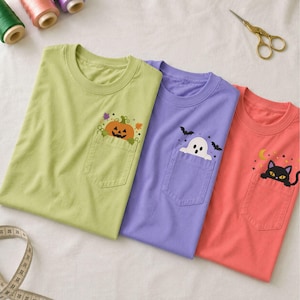 May include: Three crew neck t-shirts with chest pockets. The light green shirt features a pumpkin graphic, the purple shirt displays a white ghost and black bats, and the coral shirt shows a black cat with a yellow crescent moon and stars.
