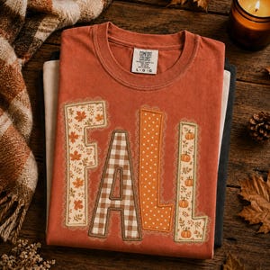 May include: A rust-colored crew neck t-shirt featuring the word FALL in large block letters. Each letter has a unique pattern: F has maple leaves, A has brown gingham, L has white polka dots, and L has pumpkins. The shirt has a Comfort Colors label.
