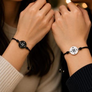 May include: Two black cord bracelets with compass rose pendants. One pendant is black with white markings, the other is silver with black markings. The bracelets are worn on wrists.