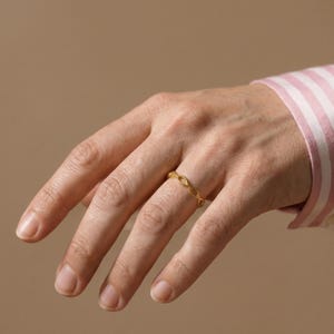 May include: A gold ring with a delicate chain-link design. The ring is worn on a finger, showcasing its intricate pattern and warm metallic hue. The ring's design is a repeating pattern of small, connected ovals.