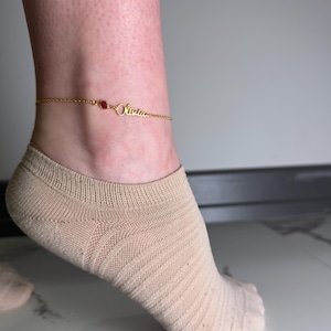 May include: Gold-colored anklet with the name "Olivia" in cursive script. The anklet features a small, round, red gemstone and is worn on an ankle. The background is a neutral gray.