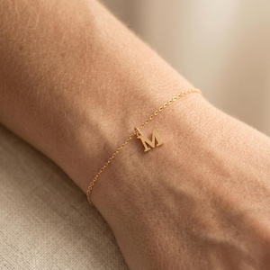 Puede incluir: Una delicada pulsera de cadena en tono dorado que presenta un único dije centrado con la letra mayúscula M. La pulsera consta de eslabones circulares finos y entrelazados que forman una banda simple y continua alrededor de la muñeca.