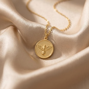 Puede incluir: Un collar dorado con un colgante redondo con un dise&ntilde;o de paloma. El colgante cuelga de una delicada cadena, sobre una tela de sat&eacute;n. La paloma est&aacute; representada con las alas extendidas y un cuerpo redondeado.