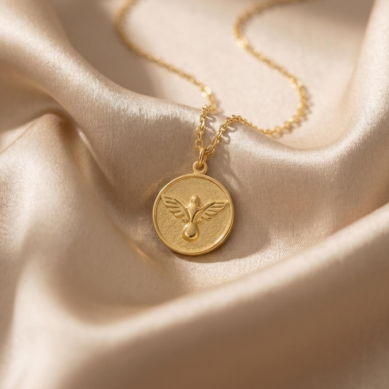 Puede incluir: Un collar dorado con un colgante redondo con un dise&ntilde;o de paloma. El colgante cuelga de una delicada cadena, sobre una tela de sat&eacute;n. La paloma est&aacute; representada con las alas extendidas y un cuerpo redondeado.