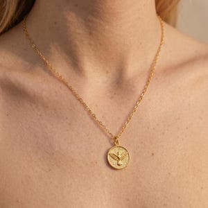 Puede incluir: Un collar dorado con un colgante circular con un dise&ntilde;o de paloma. El colgante y la cadena son de un material met&aacute;lico brillante. La paloma tiene las alas extendidas sobre un fondo texturizado.
