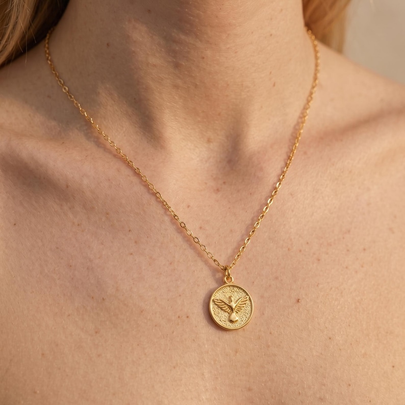 Puede incluir: Un collar dorado con un colgante circular con un dise&ntilde;o de paloma. El colgante y la cadena son de un material met&aacute;lico brillante. La paloma tiene las alas extendidas sobre un fondo texturizado.
