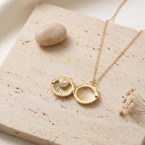 May include: A gold-colored seashell pendant necklace with a small pearl inside. The seashell locket is open, revealing the pearl and a light blue interior. The necklace is on a beige surface with a small stone and dried flowers.