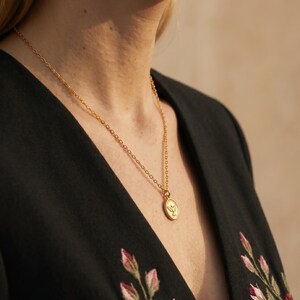 Puede incluir: Un collar dorado con un colgante con un dise&ntilde;o de hoja. El collar se lleva sobre una blusa negra con bordados florales. El colgante es ovalado y cuelga de una delicada cadena.