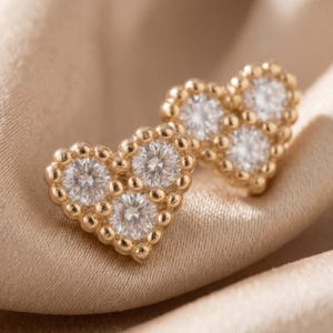 Può includere: Un paio di orecchini a bottone a forma di cuore color oro. Ogni orecchino presenta tre pietre preziose rotonde e trasparenti ed è circondato da un bordo di perline. Gli orecchini sono esposti su un tessuto beige satinato.
