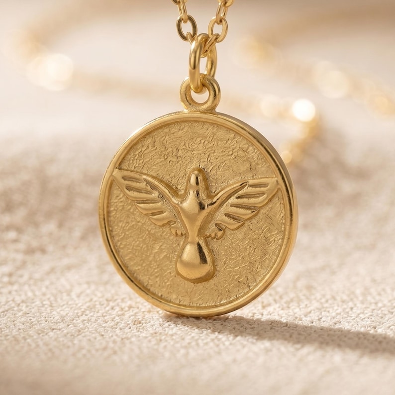 Puede incluir: Un collar con colgante dorado. El colgante circular presenta una paloma con las alas extendidas, sobre un fondo texturizado. La cadena del collar tambi&eacute;n es dorada. El dise&ntilde;o es detallado y elegante, adecuado para el uso diario o ocasiones especiales.