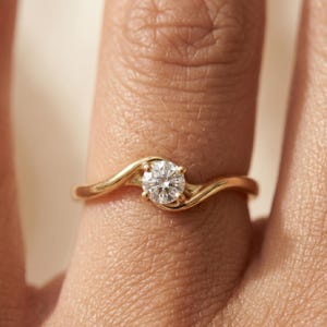 May include: A gold engagement ring with a single, round, clear diamond. The ring's band has a flowing, wave-like design. The ring is shown on a finger, with the focus on the ring's details and sparkle.