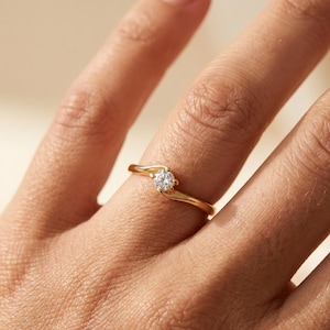 Puede incluir: Un anillo de oro con una piedra preciosa transparente de talla brillante redonda engastada en una banda de estilo bypass. La banda de metal tiene un acabado liso y pulido que se curva alrededor de la piedra central, creando un diseño fluido.