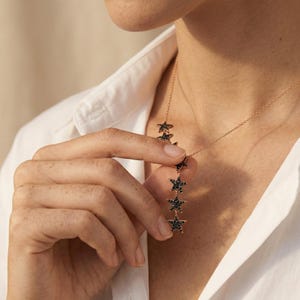 May include: A gold-toned necklace featuring a series of star pendants with black gemstone accents. The necklace is displayed against a white shirt, highlighting the contrast between the jewelry and the fabric.