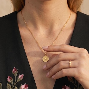 Puede incluir: Un collar dorado con un colgante redondo con un dise&ntilde;o de p&aacute;jaro. El colgante cuelga de una delicada cadena. Tambi&eacute;n se ve un anillo dorado con una peque&ntilde;a bola. El fondo incluye una blusa negra con bordados florales.