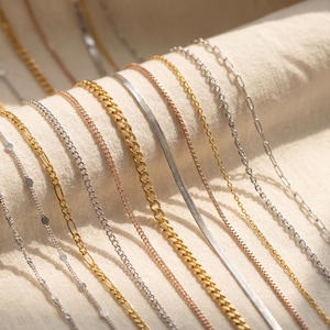 Op de afbeelding: Een verzameling van diverse metalen kettingen die parallel zijn gerangschikt, waaronder zilverkleurige, goudkleurige en roségoudkleurige stijlen. Het assortiment bevat diverse schakelontwerpen zoals gourmet-, slangen-, anker- en figaroschakels in verschillende breedtes.