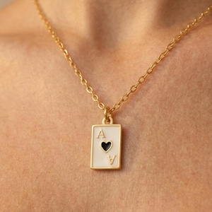 Könnte beinhalten: Eine goldfarbene Halskette mit einem rechteckigen Anhänger in Form einer Herz-Ass-Spielkarte. Der Anhänger hat eine weiße Emaille-Vorderseite mit einem schwarzen Herzsymbol und dem Buchstaben A in der oberen linken und unteren rechten Ecke.