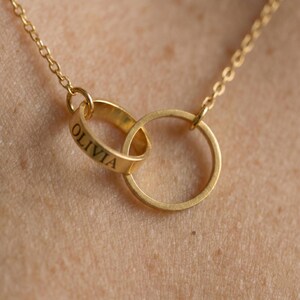 Op de afbeelding: Een goudkleurige ketting met twee in elkaar grijpende cirkelvormige ringen die aan een fijne ketting hangen. Een van de ringen is gegraveerd met de naam OLIVIA in een duidelijk, donker, schreefloos lettertype. Het metaal heeft een gladde afwerking.