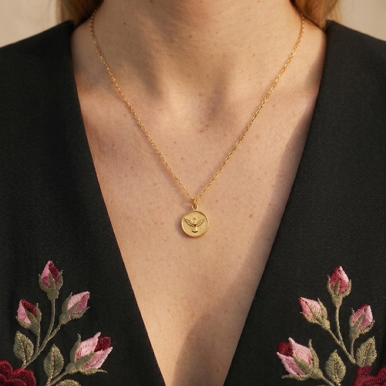 Puede incluir: Un collar dorado con un colgante redondo con un dise&ntilde;o de p&aacute;jaro. El collar se lleva sobre una blusa negra con bordados florales en tonos rosa y rojo. El colgante es circular y tiene un emblema detallado de un p&aacute;jaro.