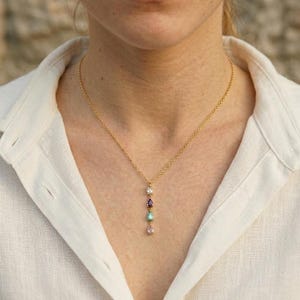 May include: A gold necklace with a pendant of four gemstones: clear, purple, teal, and pink. The necklace is worn against a white collared shirt.