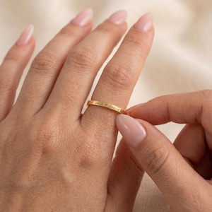 Op de afbeelding: Een dunne goudkleurige ring met een gladde, gepolijste afwerking. De buitenkant van de ring is gegraveerd met de tekst "Jack ♥ Gracie" in een duidelijk, donker lettertype, horizontaal geplaatst in het midden van de metalen band.